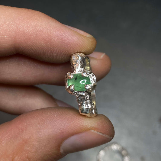 Silver ring with an emerald gemstone held between fingers against a neutral background. Daras Space